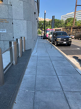 A picture of a sidewalk. There are bollards and the side of a building running from the foreground to the background of the picture on the left. Automobiles are parked on the side of the street to the right. There is a sidewalk running through the middle of the picture from the foreground to the background.